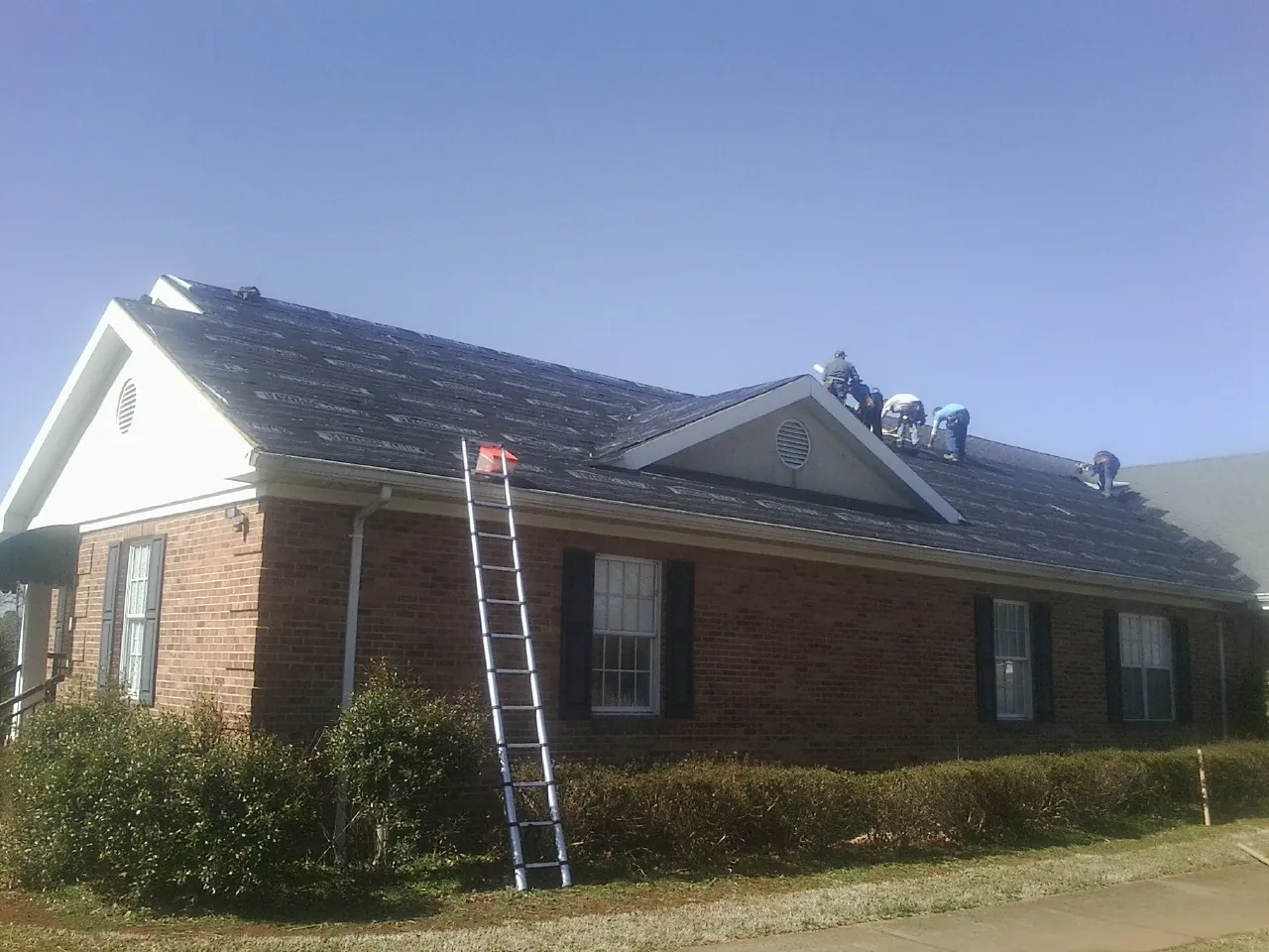 Crew tearing shingles off a brick ranch roof with a ladder set — Emergency Roof Repair work by our crew in Pleasant Shade