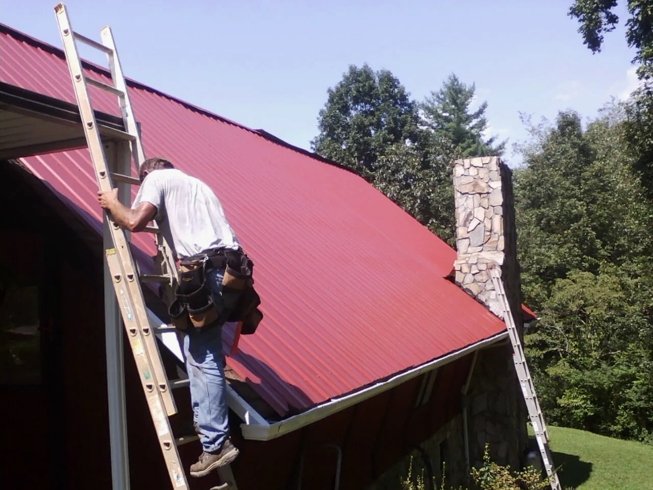 Roofer on a ladder beside a red metal roof and stone chimney — Soffit & Fascia Repair by Roofing Brothers in Pleasant Shade